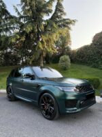 Dark green Range Rover SUV parked on a driveway with tall trees and a trimmed hedge in background.