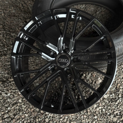 Glossy black Audi alloy wheel with a multi-spoke design resting on gravel surface.