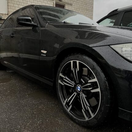 Close-up of a black BMW sedan parked in the rain, focusing on the front left wheel with a distinctive alloy rim and BMW emblem, raindrops on the body.