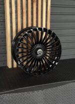 Glossy black Mercedes-Benz alloy wheel with dozens of spokes, resting against a wooden slat backdrop, center cap visible.
