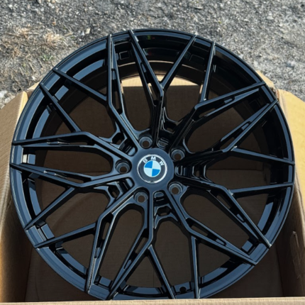 Black multi‑spoke BMW alloy wheel resting in a cardboard box on a gravel surface behind it, showing a glossy finish and center BMW emblem.