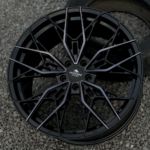 Black multi-spoke alloy wheel with an intricate star-like pattern and a Forgestar Wheels center cap on a gravel surface