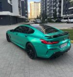 Teal BMW sports car (M4) parked on a paved residential street with modern apartment buildings in the background.