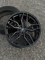 Black glossy multi-spoke alloy car wheel resting on gravel with a tire in the background, center cap visible.