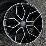 Close-up of a black alloy wheel with angular white-edged spokes on a gravel surface on a vehicle rim edge visible from the side.