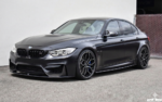Black BMW M4 sports sedan parked on pavement, showing custom multi-spoke wheels and blue brake calipers.