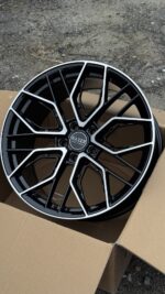 Black and silver multi-spoke alloy wheel placed in a cardboard box on a gravel surface; center cap reads HAVER.