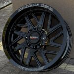 Glossy black multi-spoke alloy wheel with a raised center cap reading 'Cosmis Devil', shown at an angle against a textured wall and paver ground.