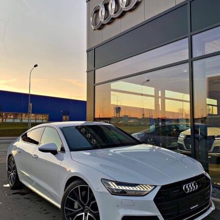 White Audi sedan parked outside an Audi dealership with the logo on the building visible at sunset.