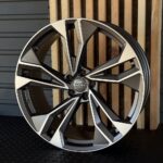 Audi alloy wheel rim with angular black and silver spokes and center cap logo against a wooden backdrop.