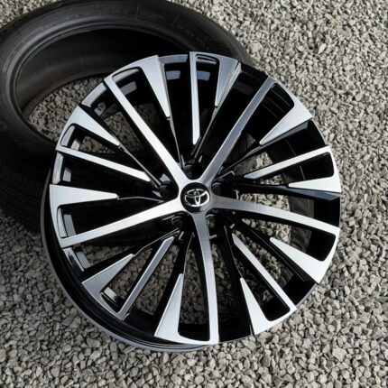 Close-up of a black and silver multi-spoke alloy wheel with Toyota center cap on a gravel surface, tire in the background.