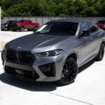 Front three-quarter view of a metallic gray BMW SUV with black multi-spoke wheels parked on a sunny dealership lot.
