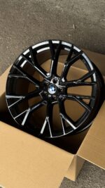 Black BMW alloy wheel with a glossy, multi-spoke design resting in a cardboard box on a gravel surface.
